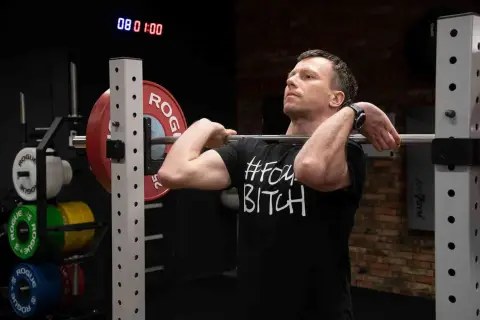 MÄÅ¼czyzna przygotowuje siÄ do przysiadu ze sztangÄ z przodu. Na koszulce widnieje napis "#FOMOBITCH".